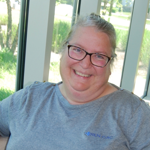 A smiling woman with gray hair and glasses stands in front of a window, wearing a gray blouse.